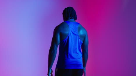 Rear-View-Studio-Portrait-Of-Male-Basketball-Player-Wearing-Team-Vest-Bouncing-Ball-Against-Colourful-Low-Key-Mixed-Lighting