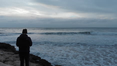 drone captures a man standing on a rocky sea shore looking out at the dramatic morning waves