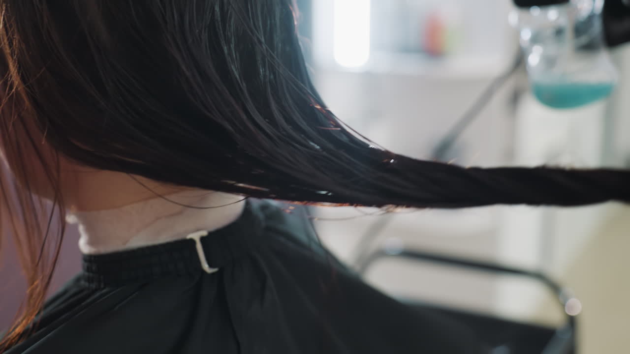 미용실에서 젖은 갈색 머리를 한 백인 여성 고객이 망토를 두르고 앉아 있고, 스타일리스트가 염색 준비를 하는 동안 젖은 머리를 빗질하고 있어요. 은은한 거울 조명과 질감, 윤기, 목, 편안한 표정이 보이는 클로즈업 세로 사진이에요.