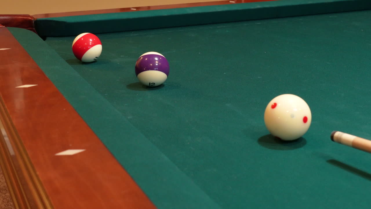 Persona che gioca a biliardo esegue un tiro combinato nella buca d'angolo con la palla 12 a strisce viola e la palla 11 rossa, usando la bilia battente mentre gioca a palla 8, primo piano della stecca in legno e del panno verde.