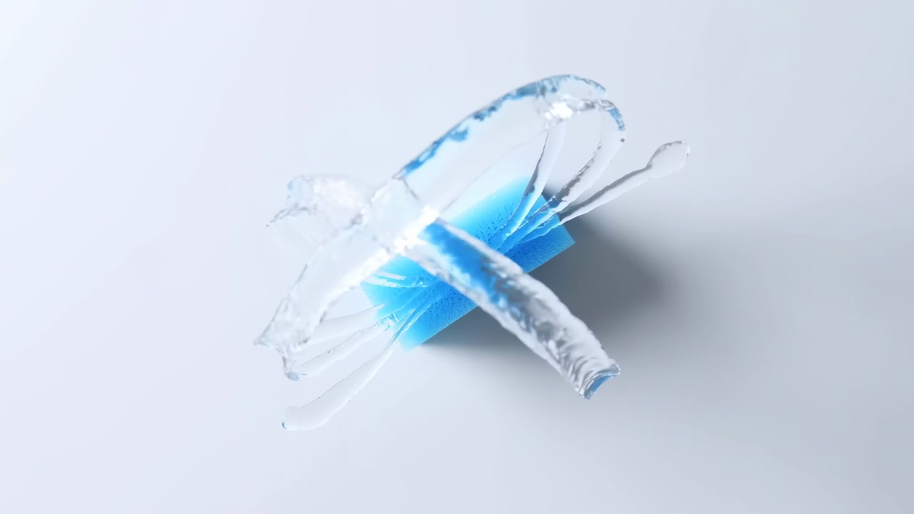 Líquido transparente se expande ao redor de uma esponja porosa azul vívida, formando fitas translúcidas arqueadas e gotículas suspensas sobre um fundo claro, enquanto a iluminação direcional projeta uma sombra cinza suave