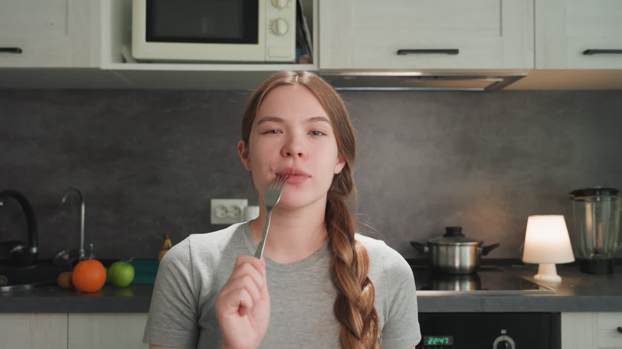 Mulher sorridente provando comida no balcão, segurando garfo, cabelo trançado, fogão e micro-ondas visíveis, luz brilhante da lâmpada, expressão brincalhona, energia de vlogger, camisa cinza casual, degustação de receita