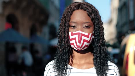 close-up view of african american woman with curly hair wearing mask looking at camera and smiling in the street