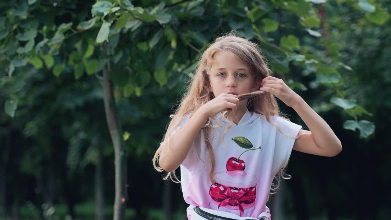 Portrait of a little girl playing with her long blond hair. Girl in the park in summer. Emotions of a child
