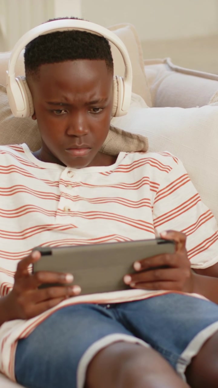Vertical video: Glancing left teen tapping gray tablet studying on couch, wearing headphones