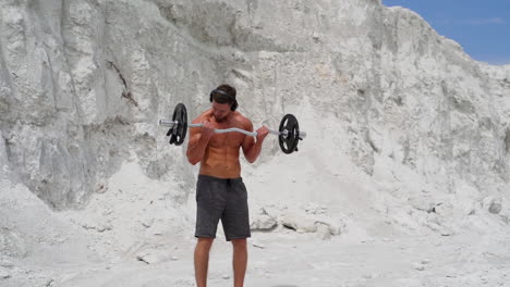 muscular bodybuilder lift barbell on white mountain background. strong young athlete with headphones doing his workout outdoors. slow motion.