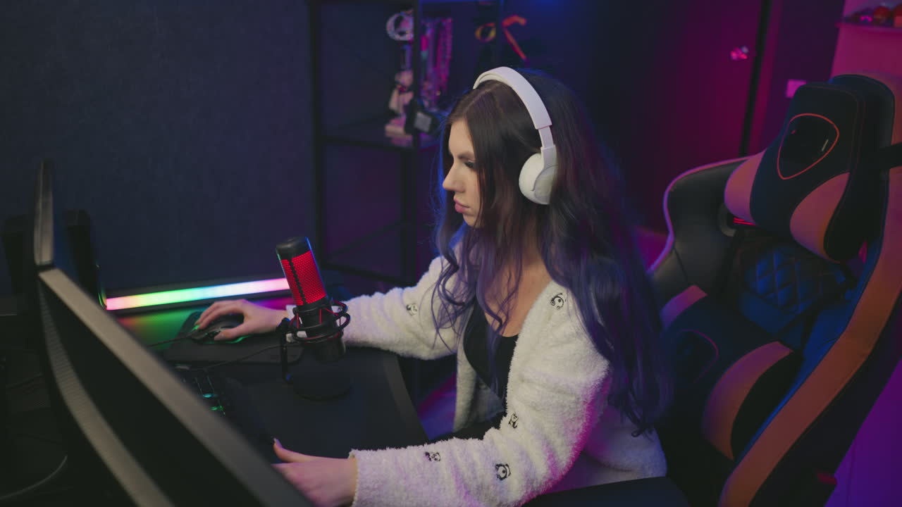 young girl wearing white headphones seated on gaming chair playing video game at desk with focused expression, colorful ambient lighting, visible microphone, and shelf in modern gaming setup
