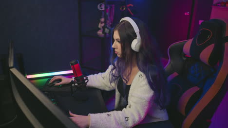 young girl wearing white headphones seated on gaming chair playing video game at desk with focused expression, colorful ambient lighting, visible microphone, and shelf in modern gaming setup