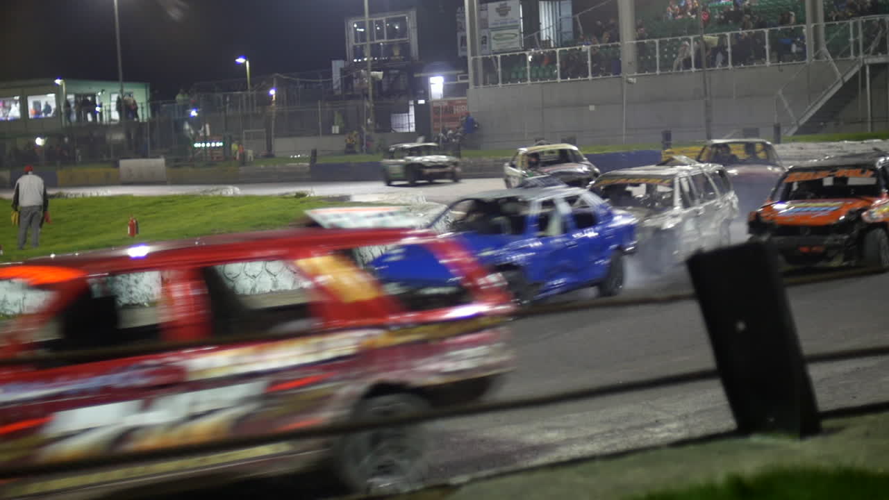 Demolition derby cars collide as they race around a corner, with smoke rising from the impact. Speeding vehicles with heavy damage on the track, spectators watch the chaotic action from the stands