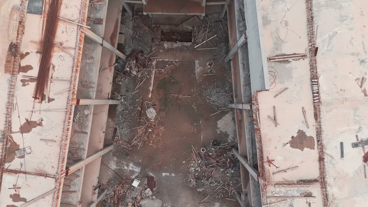 Aerial drone view of a massive, unfinished commercial building complex. Decaying concrete skeleton with unused materials and weeds, symbolizing economic recession and halted development