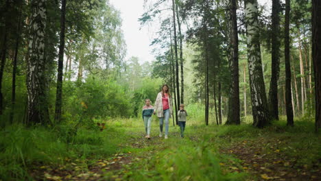 young mother adjusts hair while walking along forest trail with daughter and son, holding daughter hand as boy focuses intently on object in hand, surrounded by tall trees and lush greenery