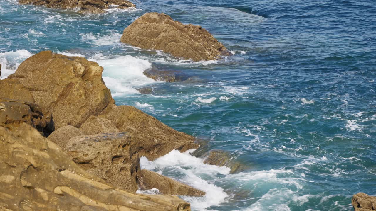 Clear blue ocean water washes over textured, sun-drenched coastal rock formations