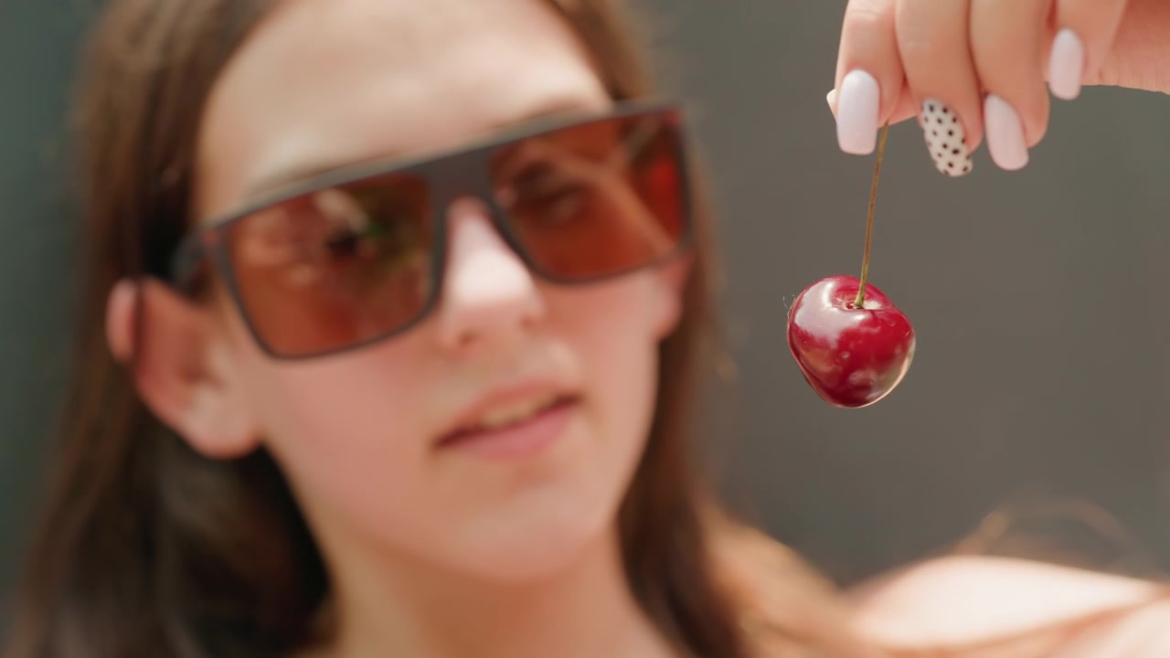 Una giovane donna con occhiali da sole tiene in mano una ciliegia con il picciolo, con il frutto a fuoco su uno sfondo semplice. I suoi capelli sono lunghi e castani. La donna si muove occasionalmente leggermente ma rimane per lo più immobile.