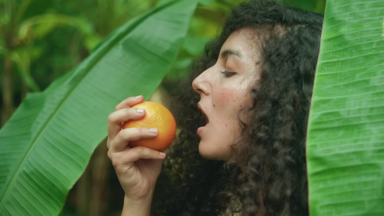 Acercamiento lento con dolly de una mujer de piel morena y cabello largo, oscuro y rizado, mordiendo una naranja fresca, rodeada de grandes hojas de plátano tropicales, evocando belleza natural y conexión sensorial