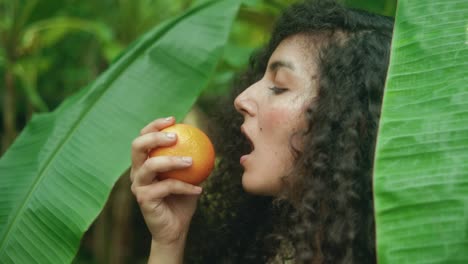 slow-motion dolly-in of woman with olive skin, and long dark curly hair biting into a fresh orange, surrounded by large tropical banana leaves, evoking natural beauty and sensory connection