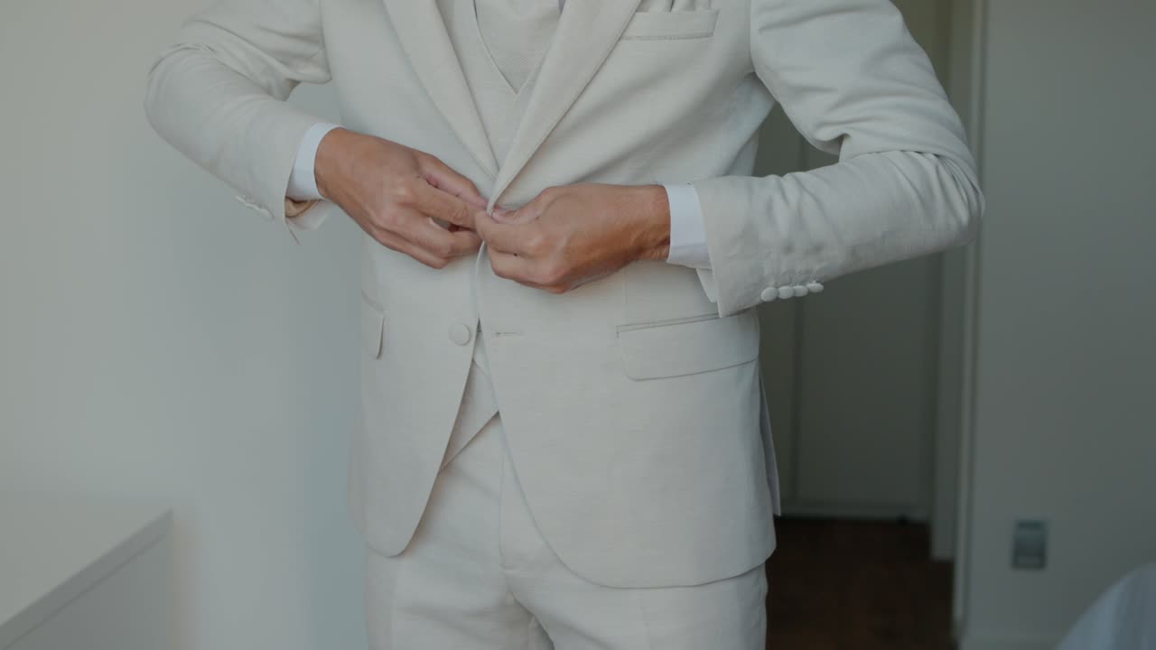 은 베이지색 코트 버튼을 고 있는 남자, 실내에서 공식적인 행사를 준비하고 있다