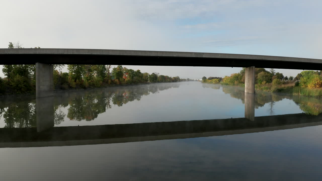 Drone flying low over a calm river with thin fog passes under a concrete bridge