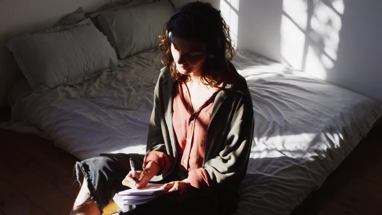 Donna pensierosa di etnia mista seduta sul letto e che scrive nella luminosa camera da letto di un cottage.