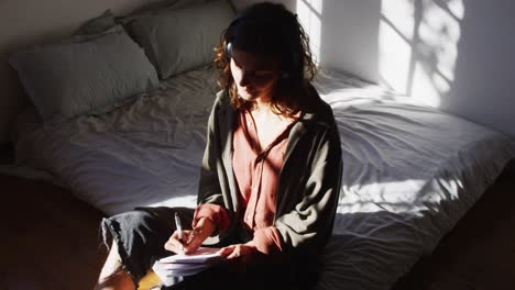 thoughtful mixed race woman sitting on bed writing in sunny cottage bedroom