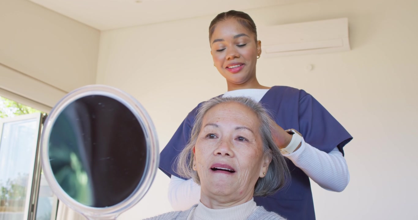 Kvinnelig pleier begynner å børste med bred børste, friserer eldre asiatisk kvinne som ser seg i speilet hjemme.