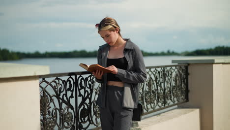 young lady in grey clothing and black handbag stands by a fence, focused on reading a book as the wind blows the pages, she turns to a new page, with trees in the background and a lamp post nearby