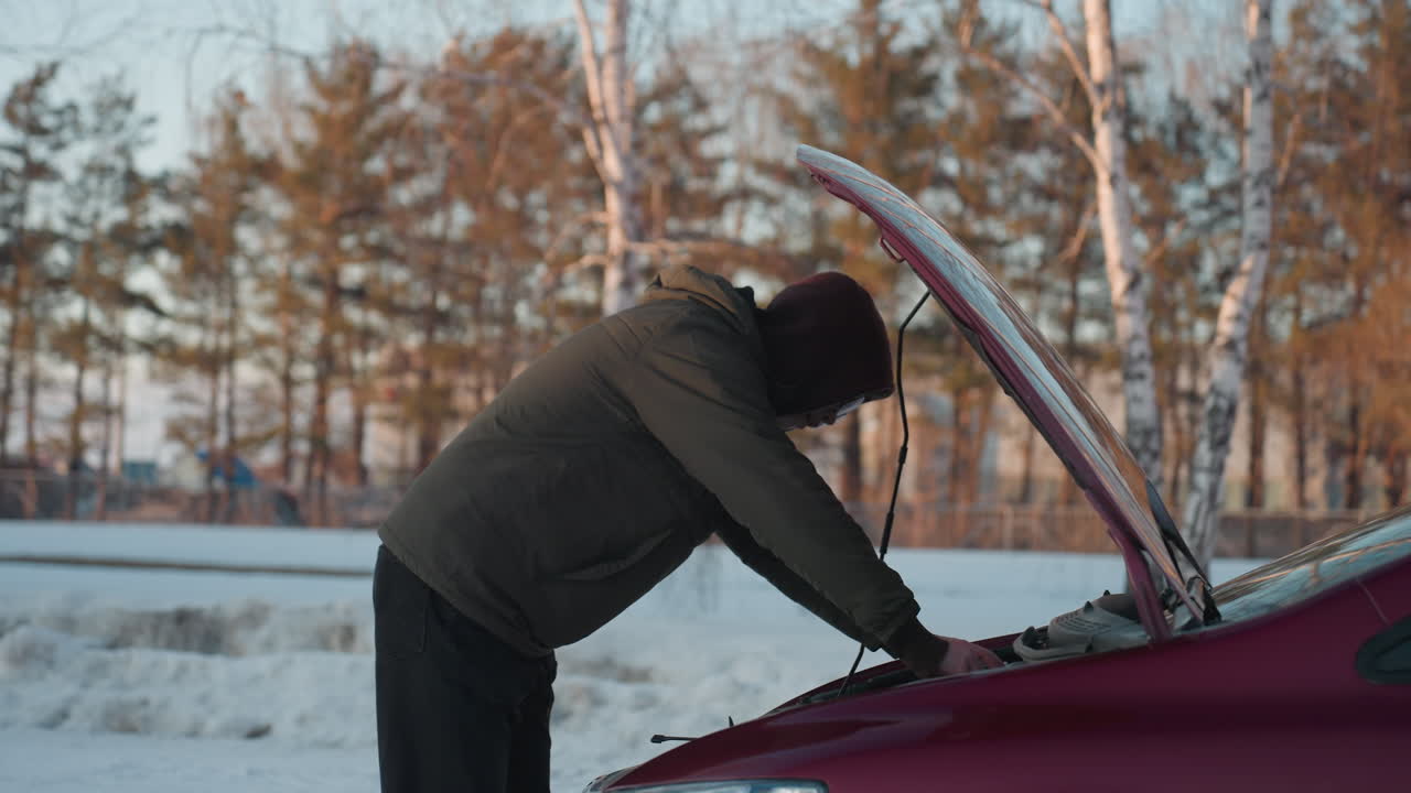 Man wearing hoodie examines car engine under raised bonnet during winter in snowy outdoor environment with trees and residential buildings in background