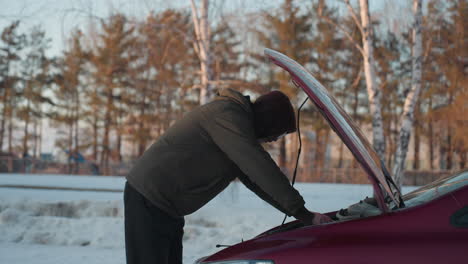 man wearing hoodie examines car engine under raised bonnet during winter in snowy outdoor environment with trees and residential buildings in background
