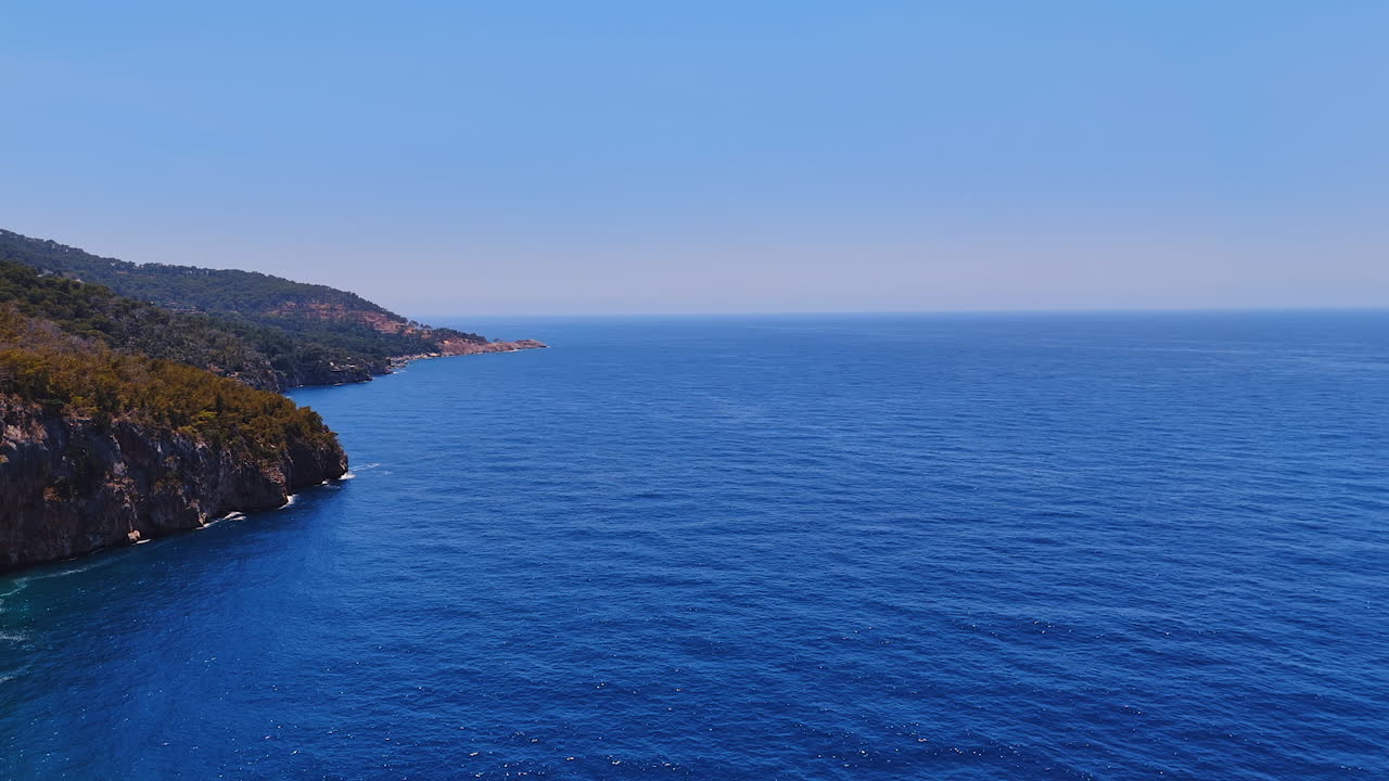 Mezzogiorno calmo sulla costa. Acque blu si estendono fino all'orizzonte, incontrando scogliere aspre e alberi verdi sotto un cielo limpido durante il giorno.