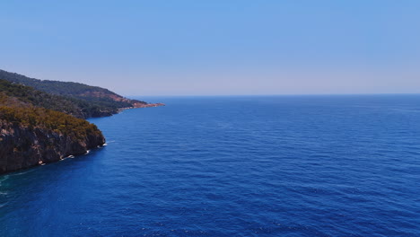 calm noon at the coastline. blue waters stretch to the horizon, meeting rugged cliffs and green trees under a clear sky in the daytime