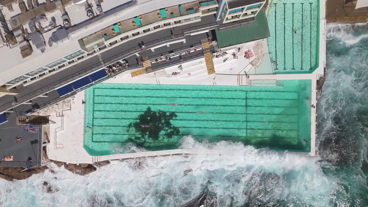 People are swimming and sunbathing at an ocean pool with ocean waves crashing on a sunny day at Bondi Beach, Sydney NSW, Australia. Aerial top view.