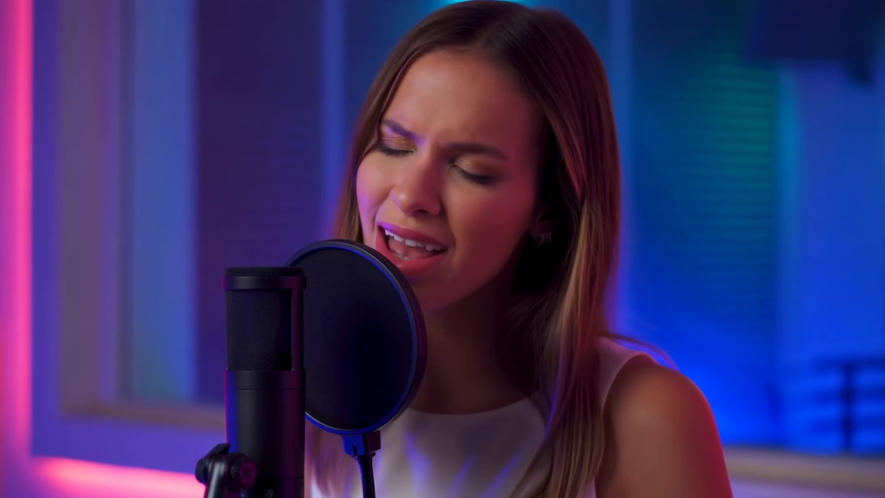 Vista frontale ravvicinata mostra una donna che canta dietro un filtro anti-pop circolare, muovendo la bocca e inclinando leggermente la testa mentre una luce colorata blu e magenta riempie lo studio