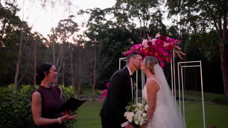 bride and groom have first kiss under pink floral arbour at outdoor wedding ceremony, 4k slow motion