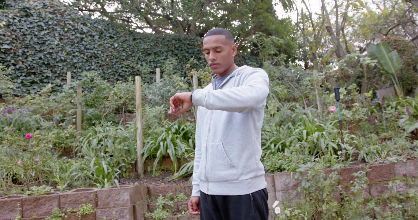 Man wearing gray hoodie and dark sweatpants in backyard glancing down, checking smartwatch for time