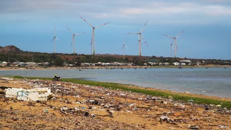 waste and dirt on sandy beach with ocean and ecological wind turbines farm for green energy during sunny day in background