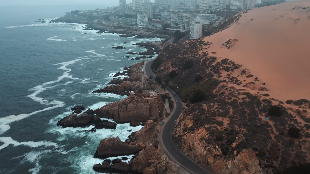 carretilla aérea en la carretera costera y dunas de arena naranja en la ladera rocosa cerca de las olas del mar que revelan edificios de concon en un día de niebla, chile
