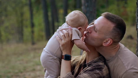 caucasian woman raises her baby over the head. mom and dad kiss their adorable infant son. blurred nature backdrop.
