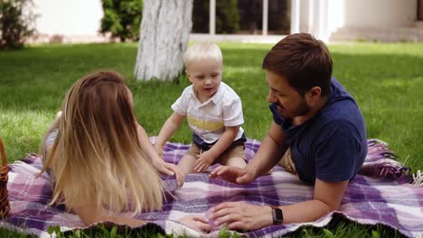 family picnic concept. young parents lying on a grass, playing with his son. have fun. child with fair hair climbs to his father's back. embraces his neck. laughing