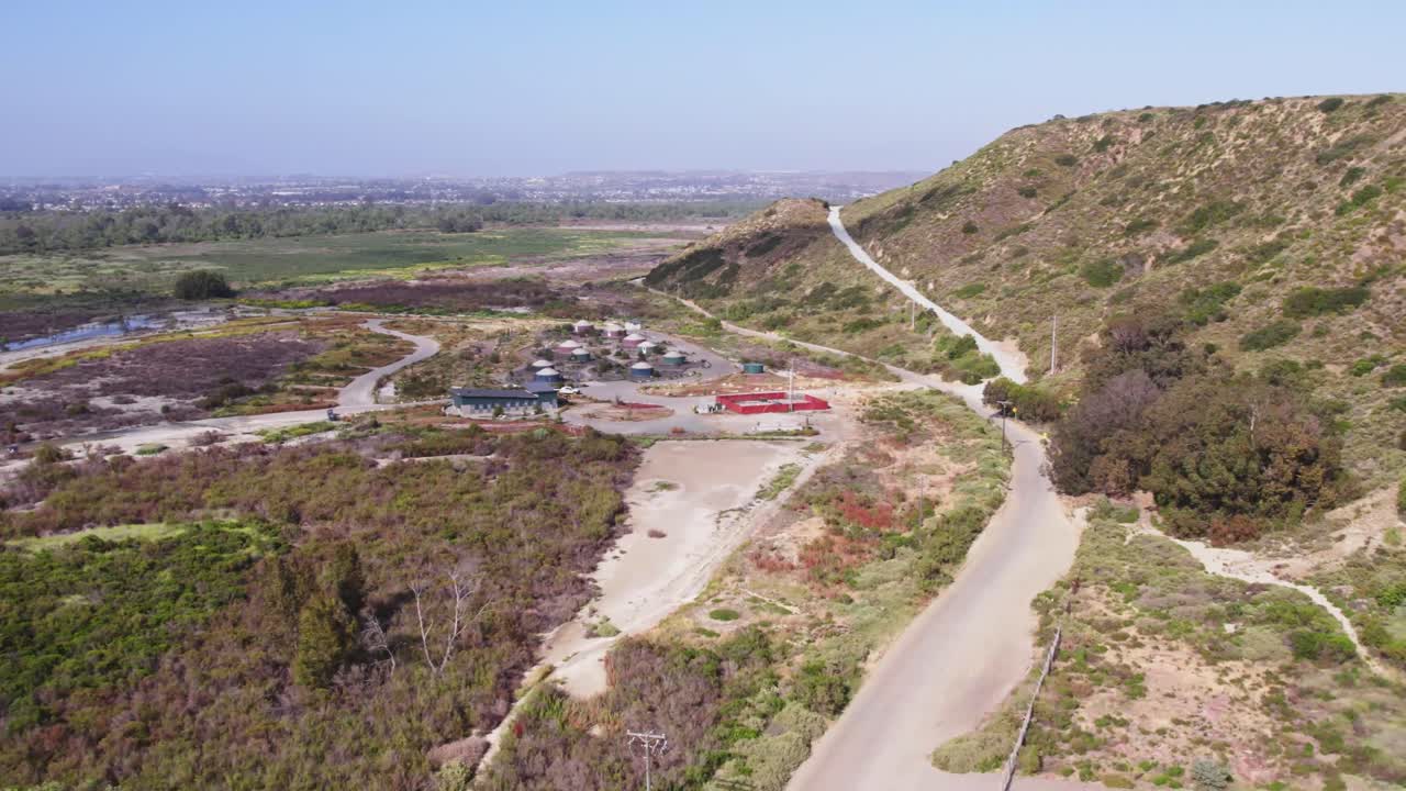 Drone aerial of a winding road passing near a settlement and along sunbaked hills near the U.S. border, showcasing rugged terrain and scattered infrastructure.