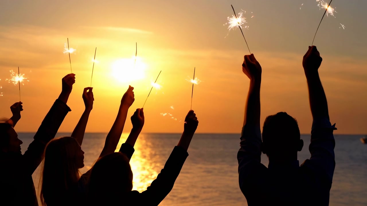 Amici in silhouette che alzano stelle filanti sull'oceano al tramonto dorato, momento celebrativo a metà sequenza