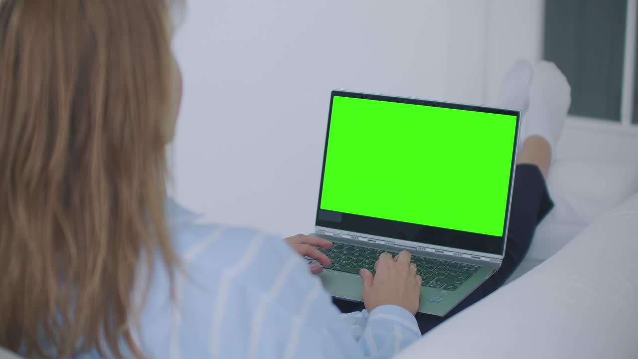 una mujer joven acostada en el sofá mira la pantalla de la computadora portátil con una pantalla verde y asiente con la cabeza. haga una videollamada