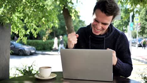 hombre emocionado celebrando el éxito en la computadora portátil sentado en la terraza del café