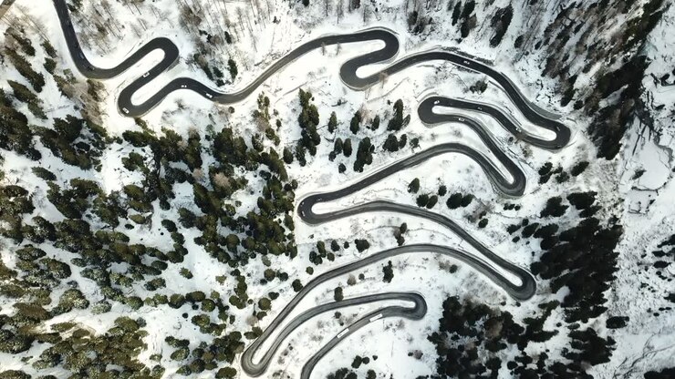 Vista aerea dall'alto che scende su una strada tortuosa attraverso il Passo del Maloja nei Grigioni, Svizzera, mostrando le curve della strada alpina in un paesaggio innevato.