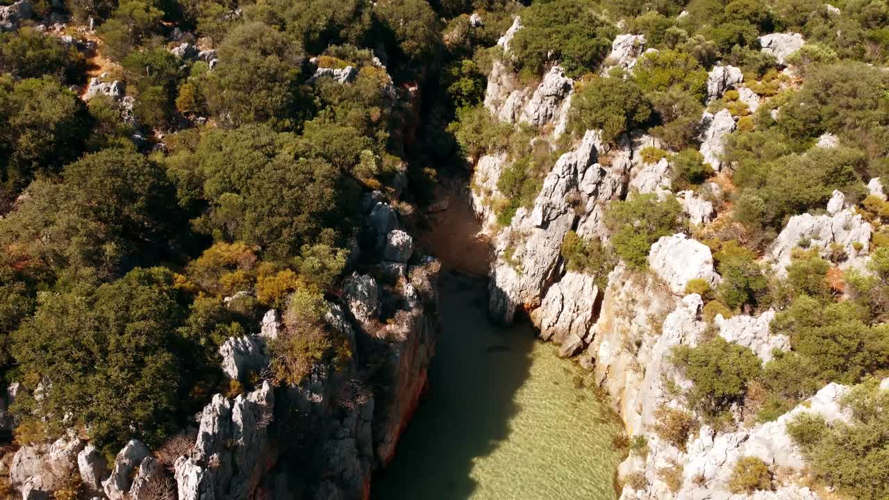 Aerial drone view showing green tinted stream flowing through narrow rocky gorge of Saklikent Canyon in Mugla Province with steep walls, dense shrubs, and bright warm daylight over rugged terrain