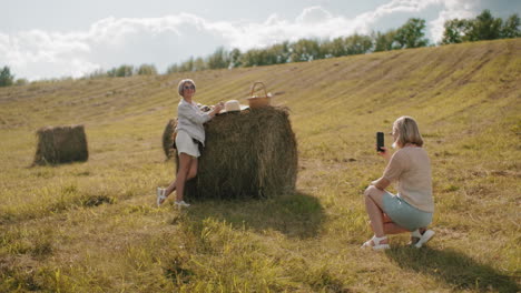 woman kneels in golden field taking photos of stylish friend leaning on hay bale with picnic setup, warm sunlight enhances rural scenery