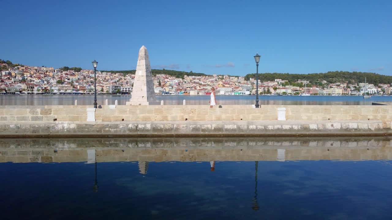 Aerial.  Young woman walking by the most famous obelisk on the De Bosset bridge. Monument in Argostoli city, capital of Kefalonia, Greece
