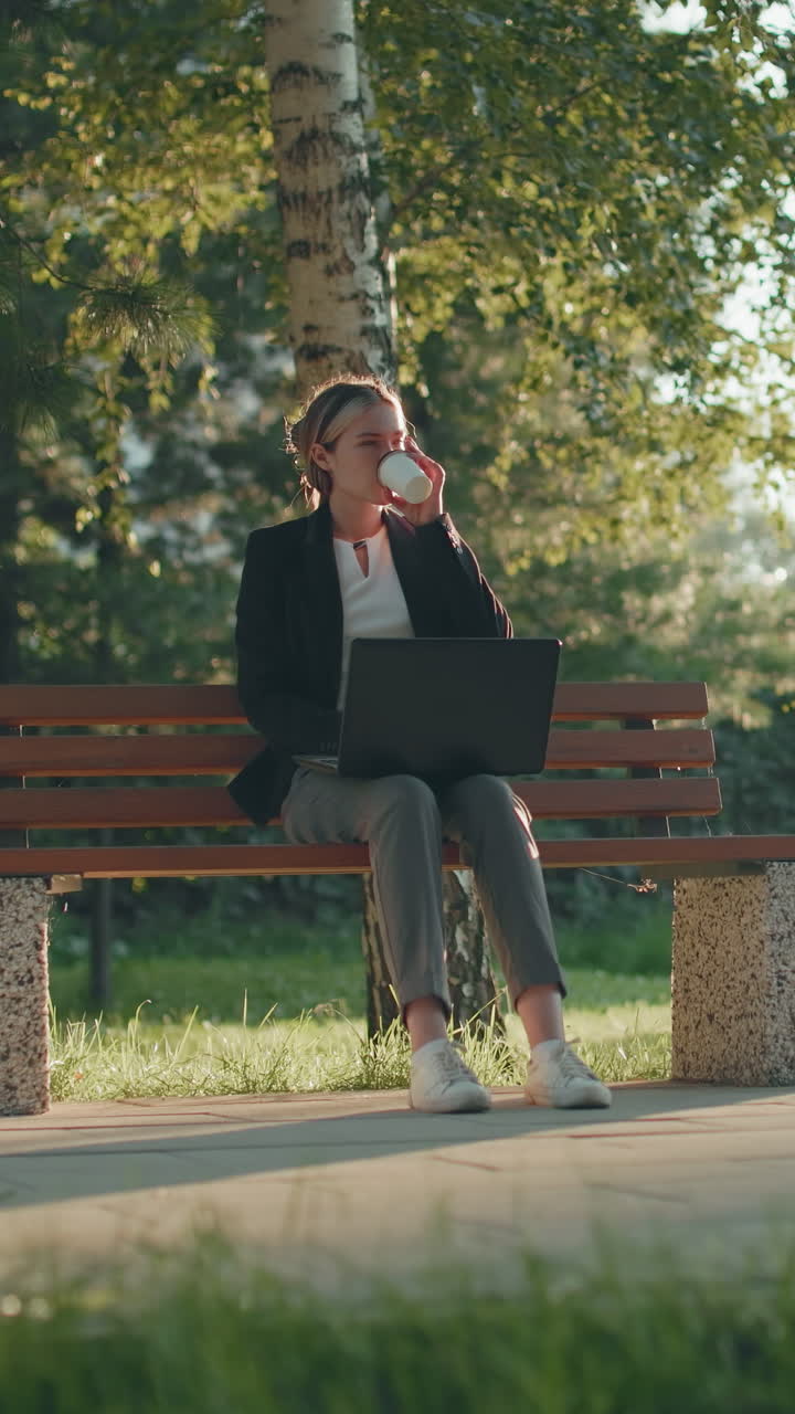 Donna d'affari seduta all'aperto in un tranquillo parco verde, che lavora al sistema e sorseggia succo con la luce del sole che filtra tra gli alberi e la recinzione di ferro sullo sfondo