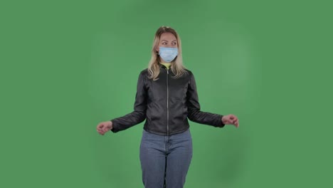 의료 보호 얼굴 마스크를 입은 아름다운 젊은 여성의 초상화는 카메라를보고 긴장하고 진정하고 명상합니다. 검은 재과 청바지를 입은 느슨한 머리카락을 가진 금발은 스튜디오에서 녹색 화면에 있습니다. 건강 보호 코로나 바이러스