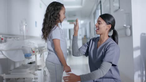 Diverse-female-nurse-and-child-patient-high-fiving-in-corridor-at-hospital,-in-slow-motion