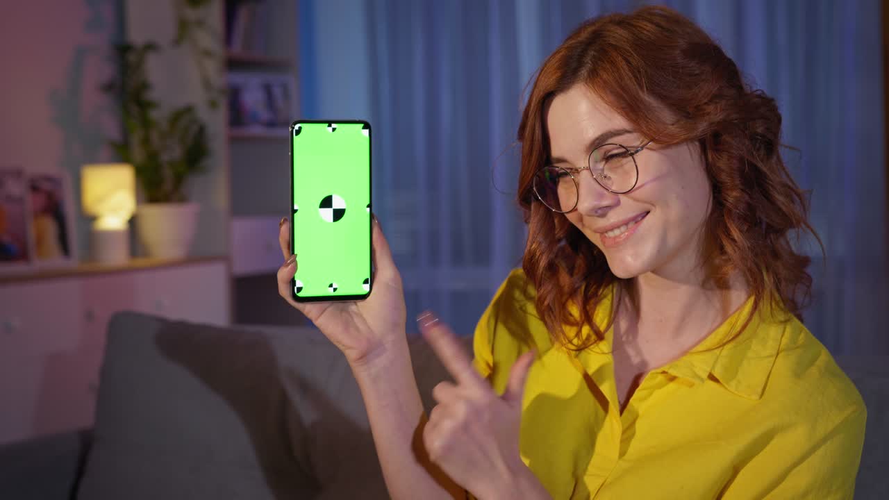 una mujer feliz que usa un navegador de internet o una red social muestra el dedo en la tecla de croma verde de su teléfono inteligente, mientras se relaja en el sofá por la noche en casa