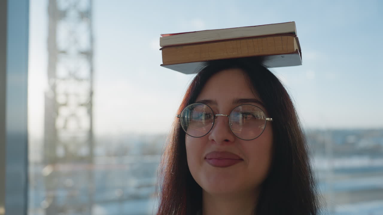 Close up of young woman turning with book on head and warm smile on face, sunlight casting soft glow through glass wall with urban structure faintly visible outside